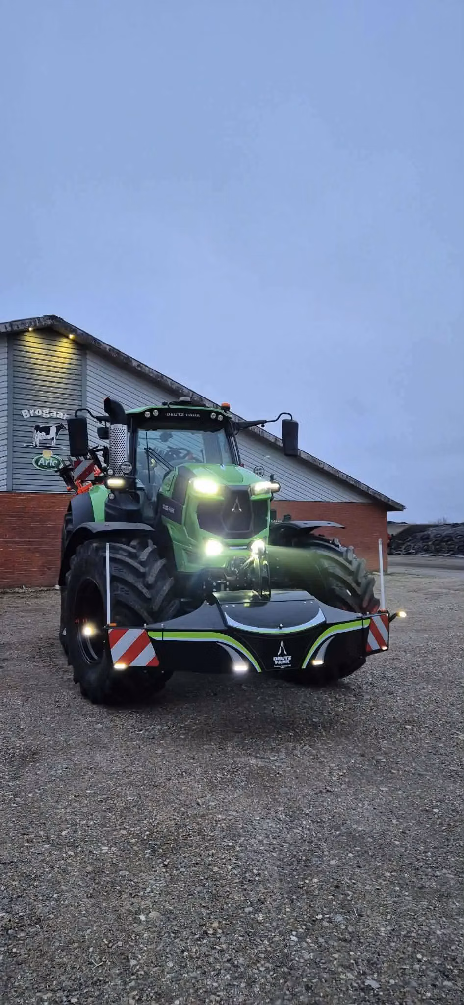 Green tractor with headlights on, fitted with a Deutz-Fahr branded front weight bumper on gravel in front of a building