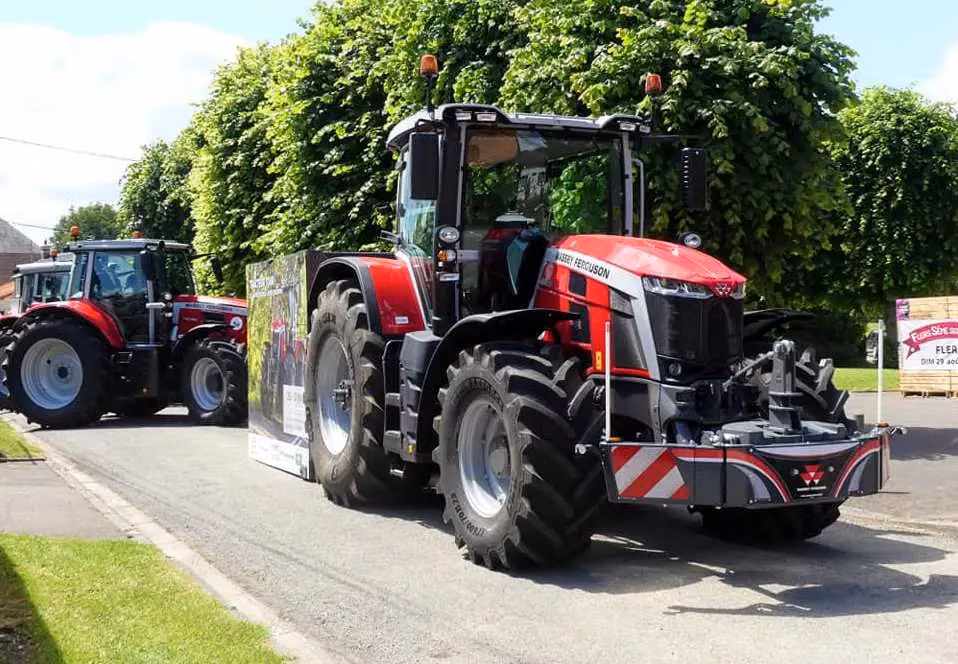 A red tractor fitted with a large front weight bumper is parked on a street with another tractor in the background