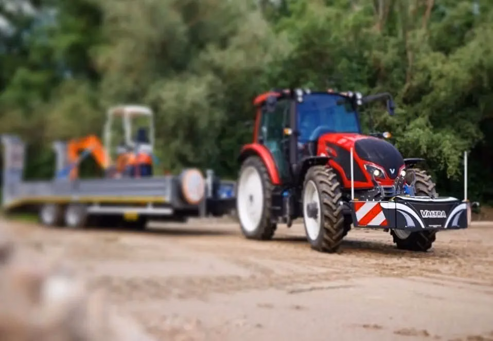 Red tractor with large front weight bumper labeled Valtra, fitted with warning stripes, parked on a dirt surface