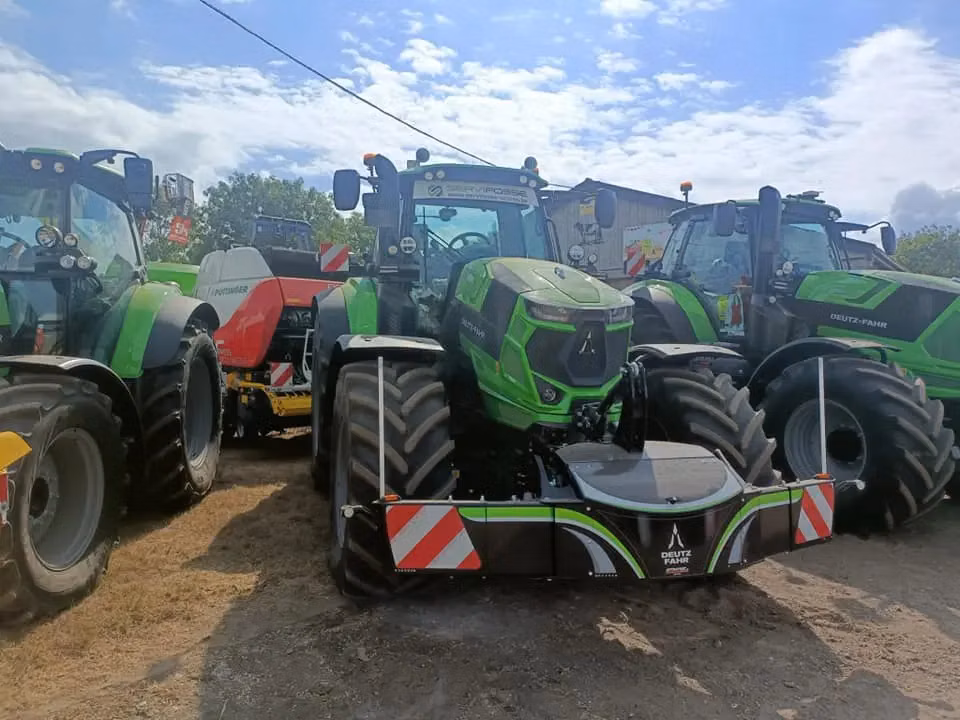 Green tractor with a large black and silver front weight bumper, parked among other tractors on a sunny day