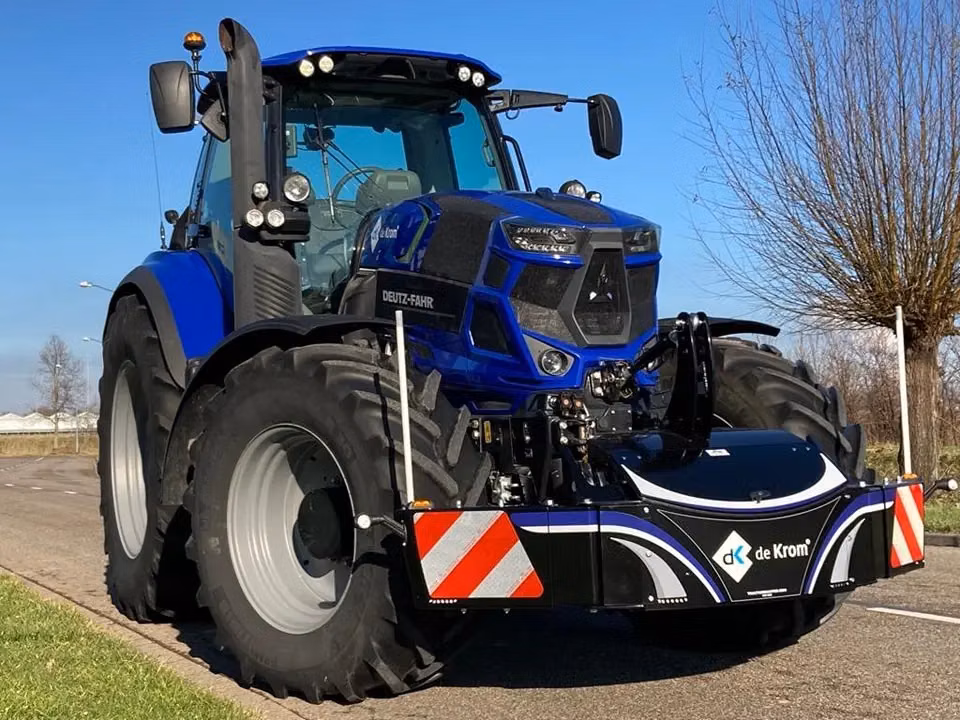Blue tractor fitted with a black front weight bumper featuring reflective red and white stripes and de Krom branding