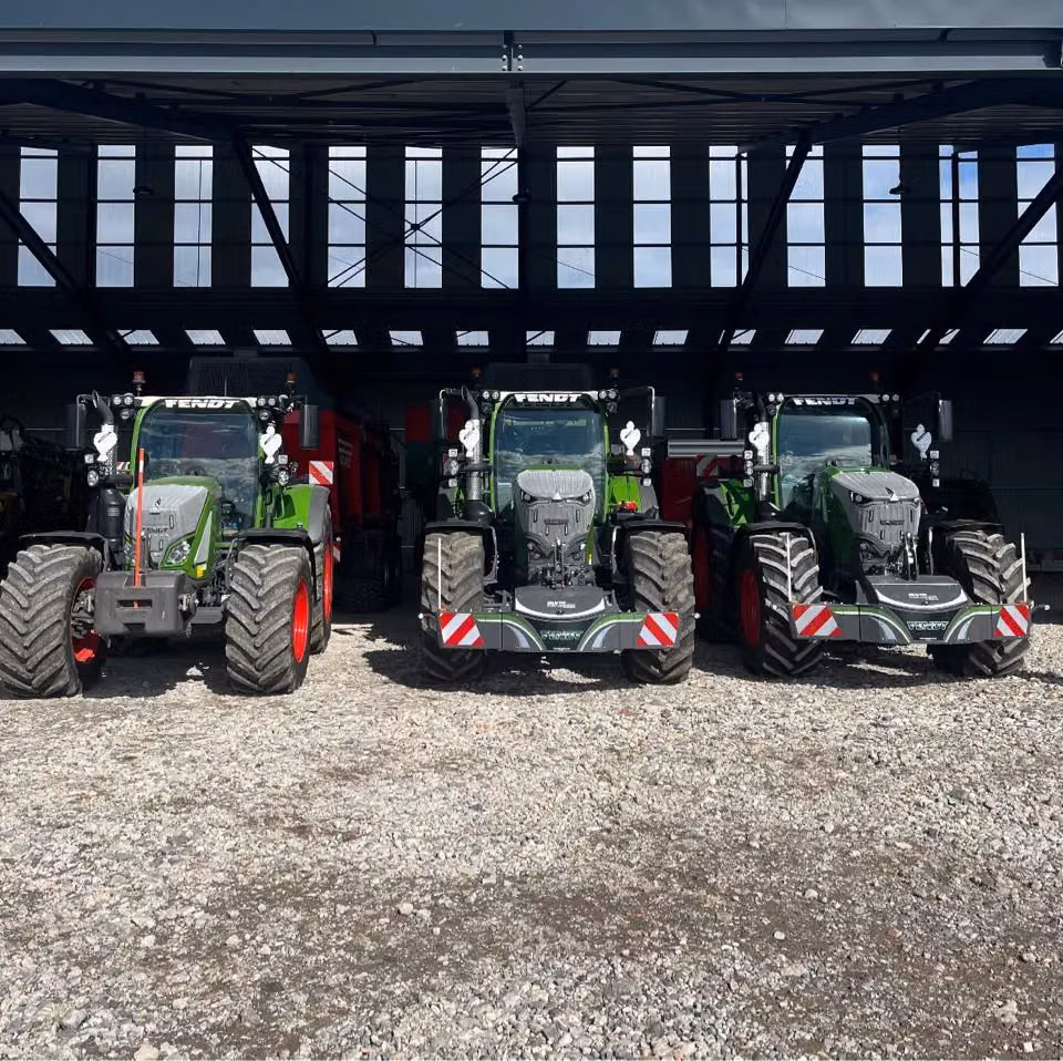 Three green tractors with front weight bumpers and red wheel rims are parked side by side on gravel under a shed