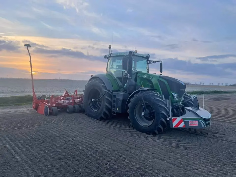 A green tractor with a front weight bumper and red agricultural attachment works in a plowed field at sunset