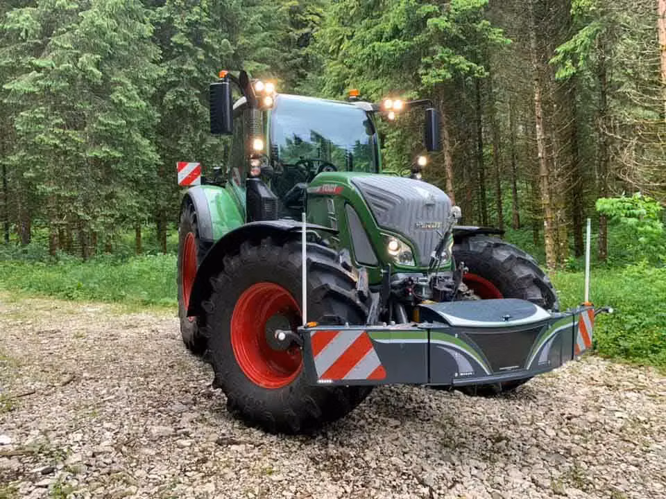 A green tractor with a front-mounted weight bumper featuring red and white warning stripes on a forest track