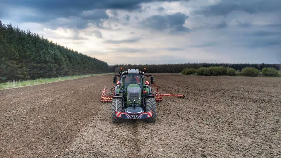 A tractor with a front weight bumper and rear attachment stands on a large plowed field under a cloudy sky