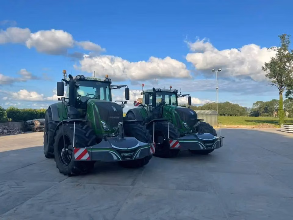 Two green tractors fitted with large front weight bumpers are parked on a concrete yard under a blue sky with clouds