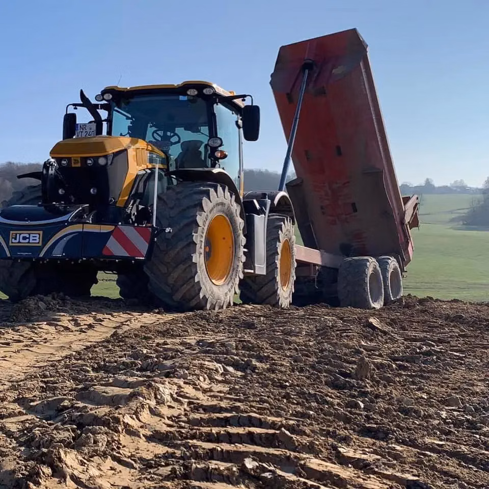 JCB tractor with a front weight bumper and LED lights attached, pulling a red tipper trailer on a muddy field