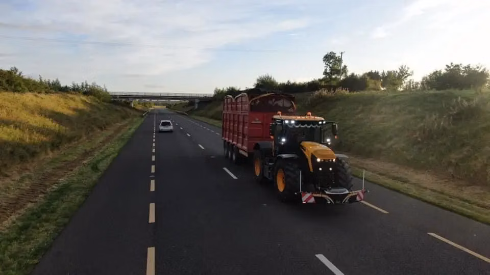 A yellow tractor fitted with a front weight bumper towing a large red trailer on a two-lane road