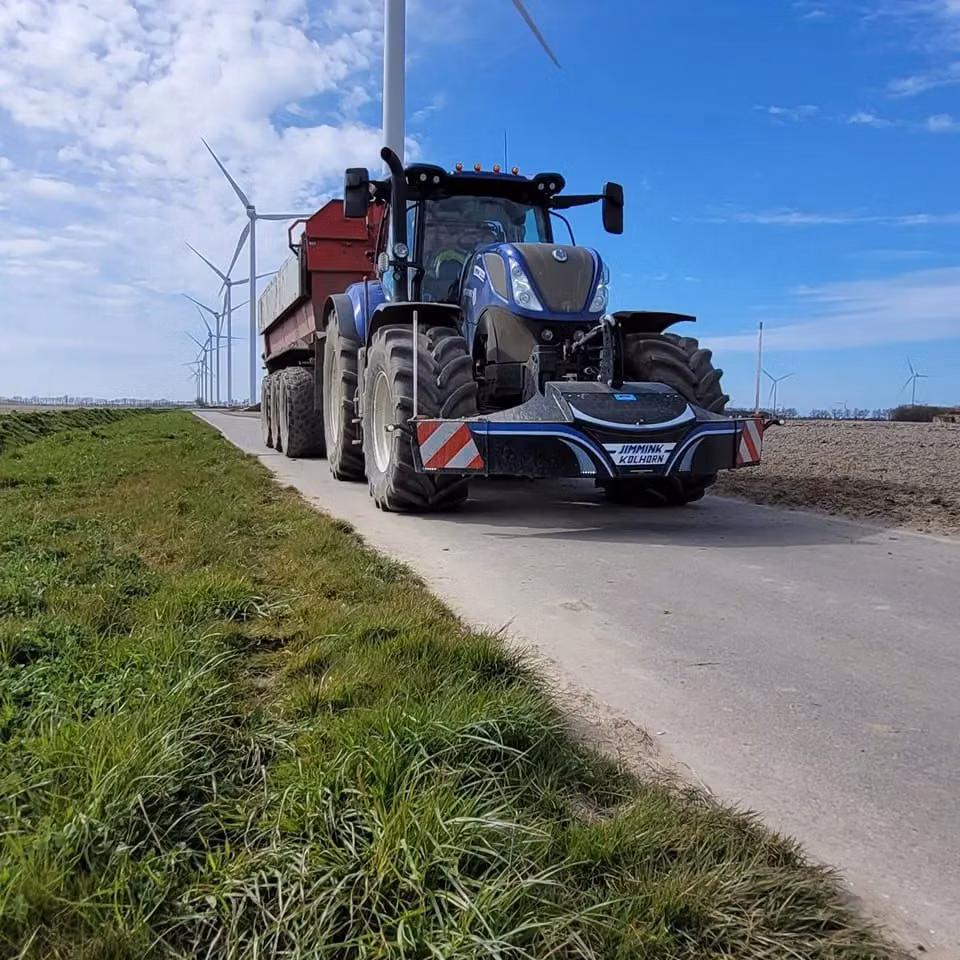 Blue tractor with a front weight bumper on a rural road, pulling a red trailer with wind turbines in the background