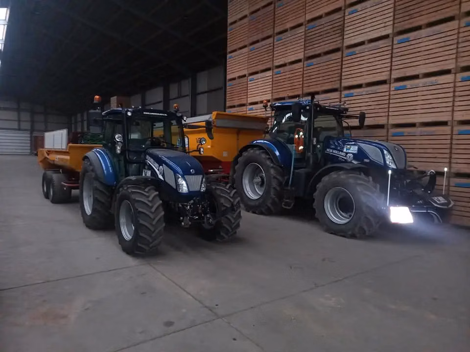 Two blue tractors with trailers are parked indoors, the tractor on the right fitted with a front weight bumper.
