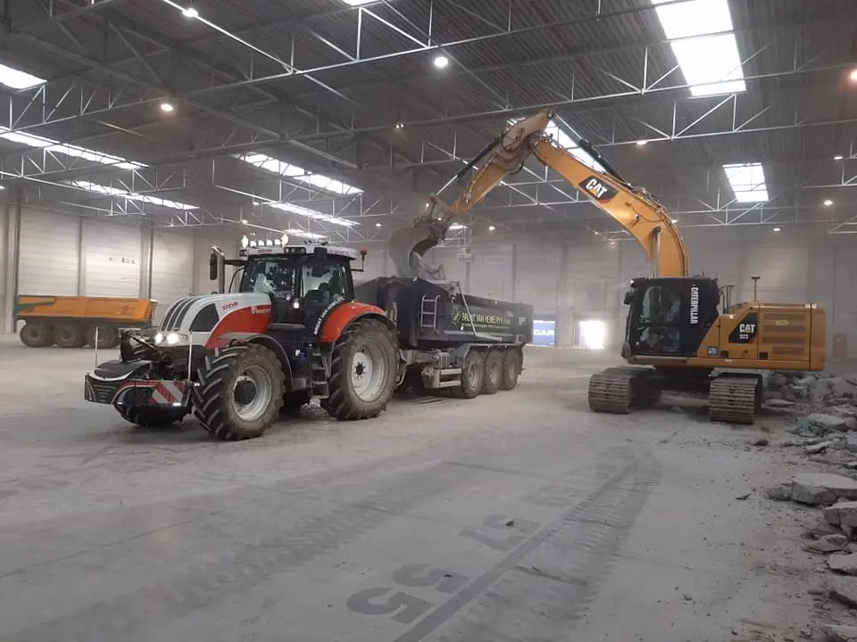 Tractor with a front weight bumper parked indoors beside an excavator loading material into a trailer