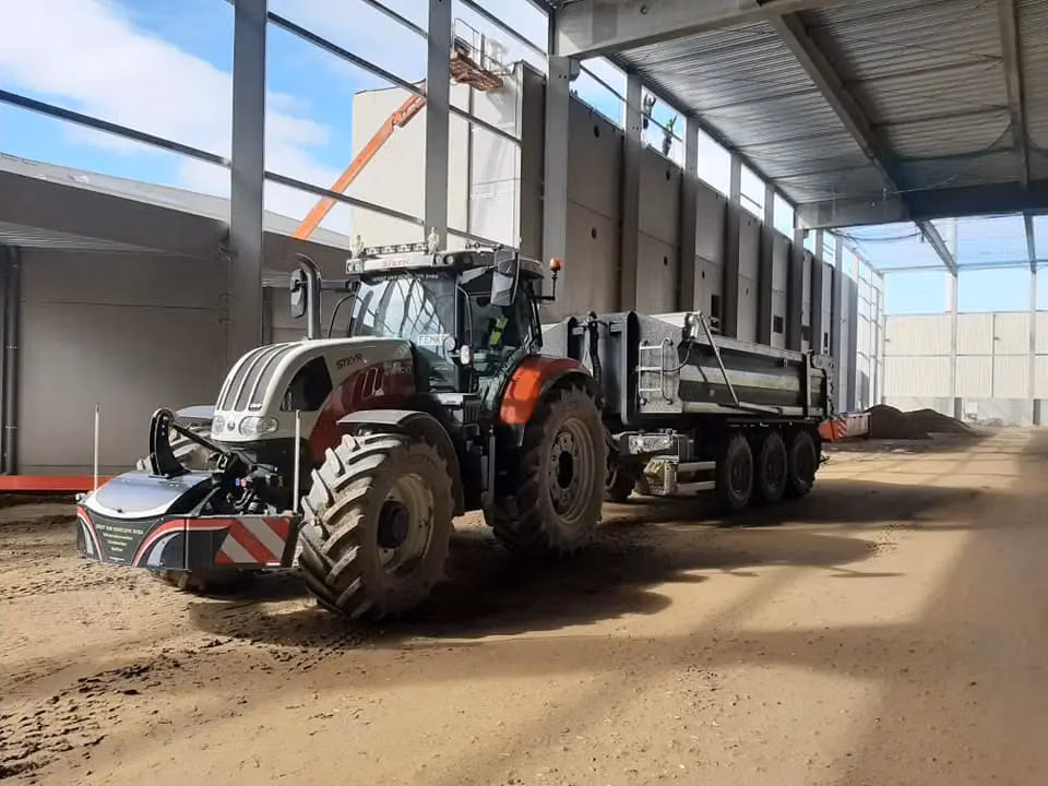 A tractor with a front weight bumper and LED lights is hitched to a large trailer inside a spacious building