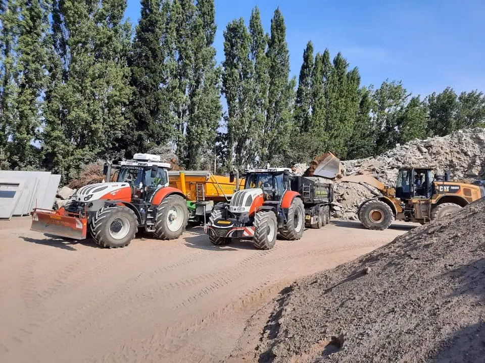 Two red and white tractors with front weight bumpers and work attachments parked on a dirt site near a loader and trees