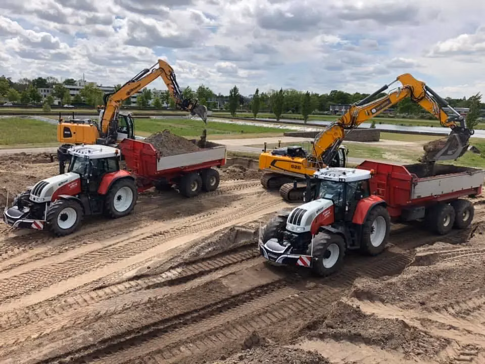 Two red and white tractors with front weight bumpers and wide marking bars are loaded with soil by excavators