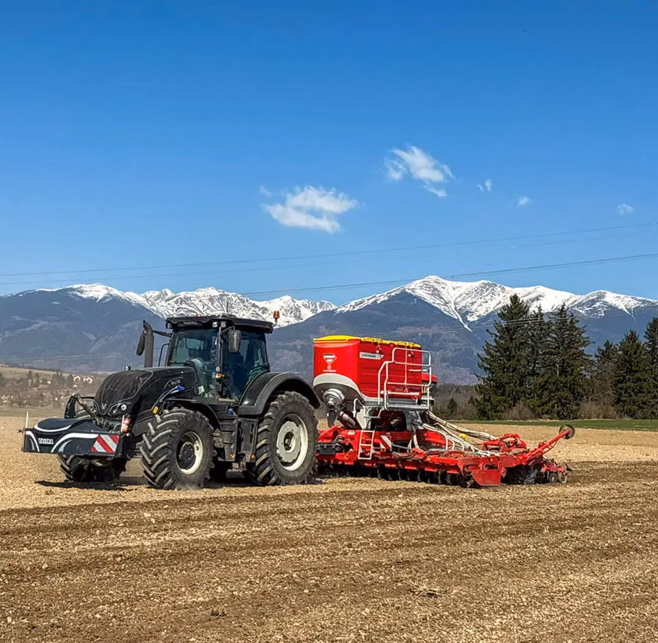 A black tractor with a front weight bumper and a red agricultural implement works in a field with snowy mountains behind