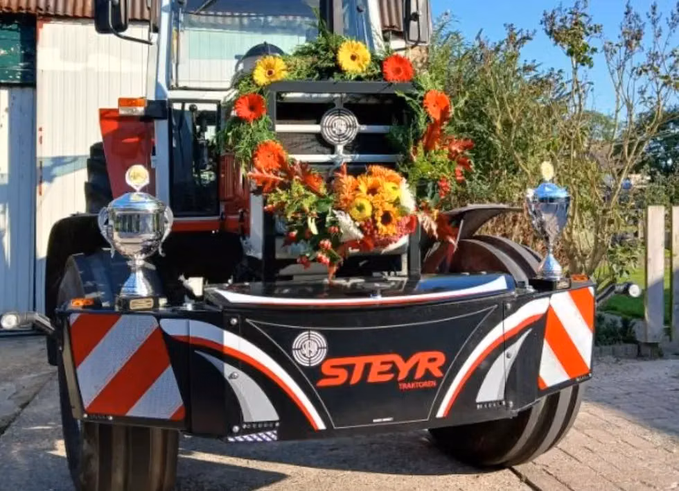 Front weight bumper with STEYR branding mounted on a tractor, decorated with flowers and flanked by two trophies